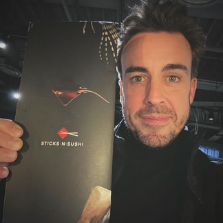 Fernando Alonso smiles holding up a Sticks n Sushi menu next to his face