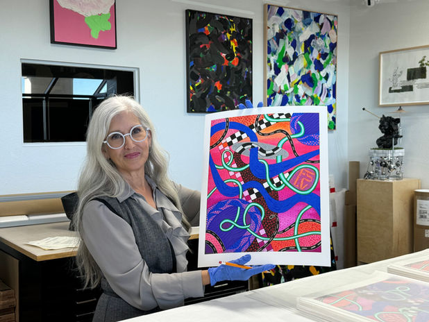 Nancy Good sitting at a desk holding up a print of her colorful painting featuring a pink background with blue, green and checkerboard swirly lines