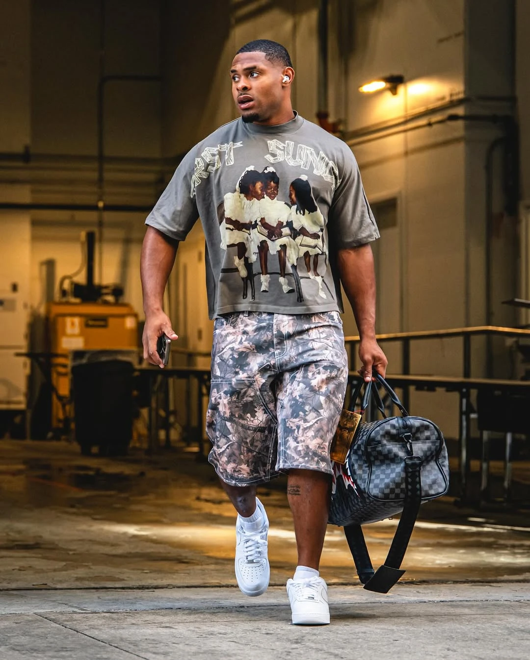 Jarquez Hunter walking out of a tunnel wearing a gray graphic T-shirt, gray camouflage pants and white sneakers and holding a checkerboard duffel