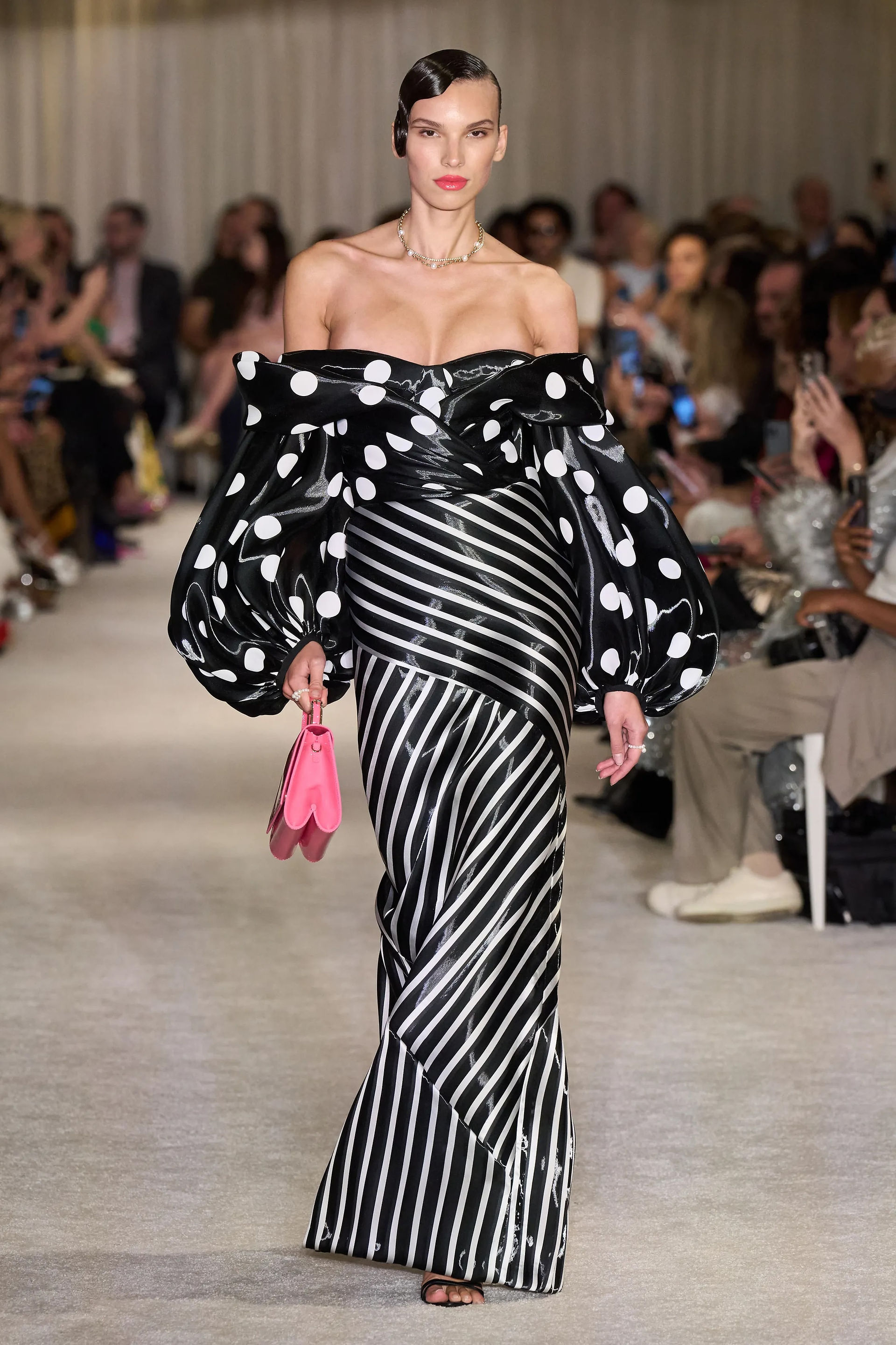 A model walking on a cream carpeted runway wearing a black polka dot off the shoulder long sleeve top and striped floor length skirt