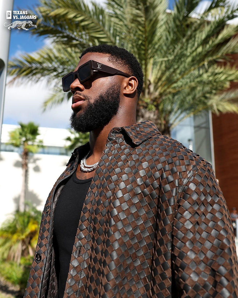 A bust shot of Josh Hines-Allen wearing Louis Vuitton shades and a Bottega Veneta brown and black woven shirt with a palm tree in the background