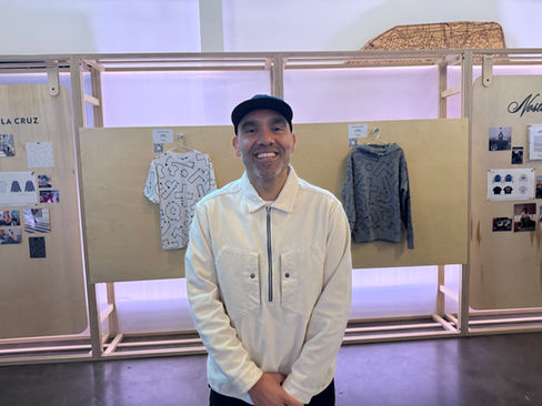 Aaron De La Cruz smiles in front of his T-shirt and hoodie on a wooden wall at the NFL Origins pop-up