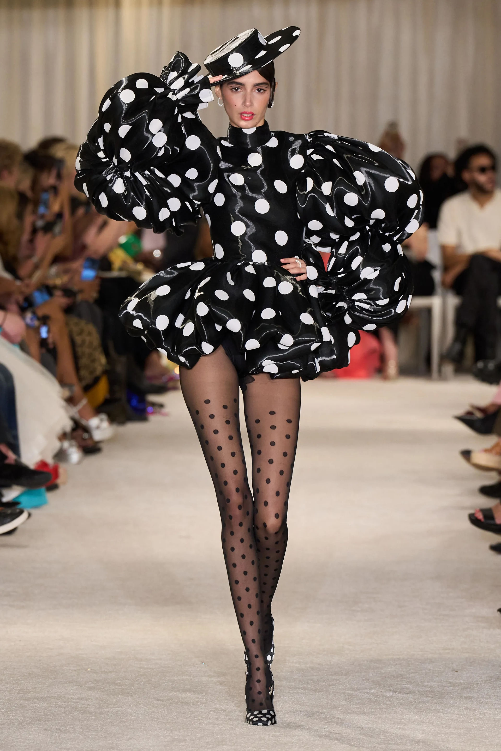 A model walking on a cream carpeted runway wearing a black polka dot bubble dress and matching hat
