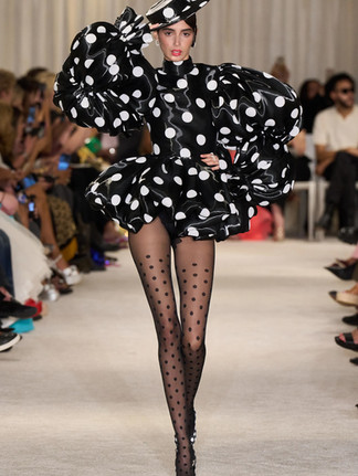 A model walking on a cream carpeted runway wearing a black polka dot bubble dress and matching hat