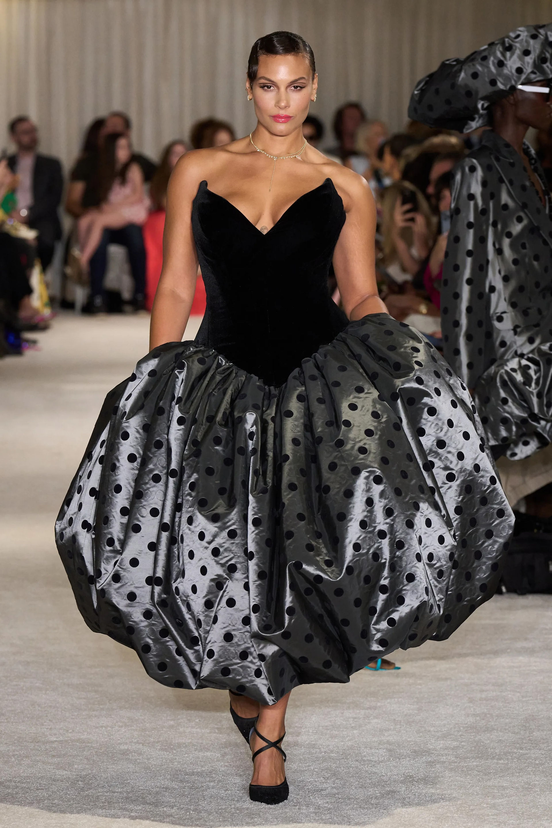 A model walking on a cream carpeted runway wearing a dress with a black sweetheart neck top and a long silver polka dot bubble skirt