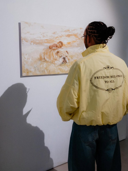 Lewis Hamilton looking at a painting of him as a kid and a bulldog in an art gallery wearing a yellow Willy Chavarria track jacket that says "Freedom Belongs To All" on the back