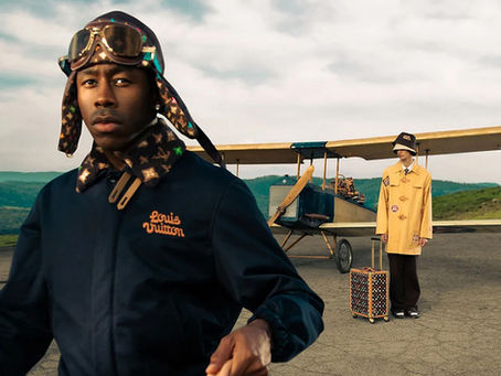 Tyler, The Creator carries a Louis Vuitton duffel bag while wearing a Louis Vuitton jacket and an aviator cap while walking away from a small plane