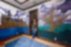 An environmental portrait of Sara Mack of the Artist Couple standing in a room surrounded by murals of Hogwarts castle and a Womping Willow tree.