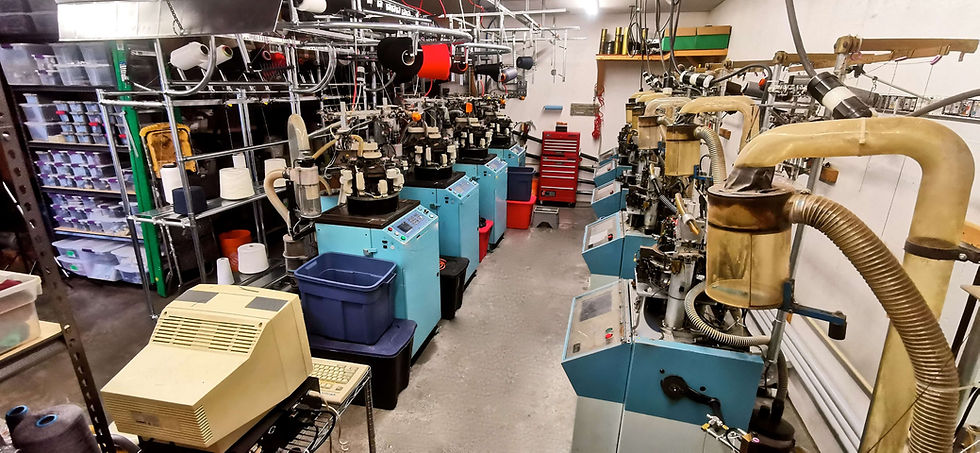 Motley Woolens knitting machines in a cluttered workshop. Shelves hold yarn spools and storage bins. A beige monitor sits in the foreground.