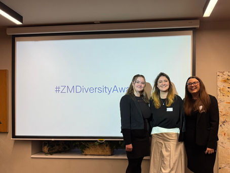Three women in black and white outfits stand smiling in front of a screen displaying #ZMDiversityAwards. The room is warmly lit.