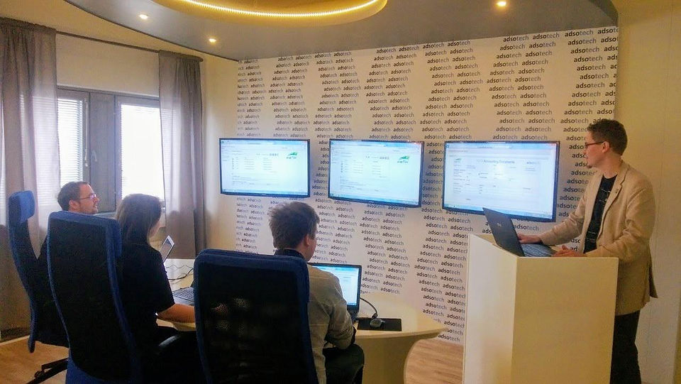 A modern Adsotech office setup with a team gathered around multiple wall-mounted screens showing SAP interfaces. One team member presents from a standing desk while others work from laptops — showcasing a collaborative, tech-driven environment.