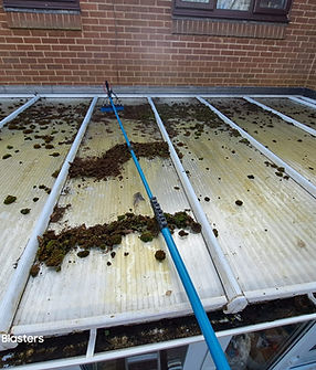 Close-up view of a conservatory roof covered with moss and dirt being cleaned using a long-handled brush. The roof panels are translucent and framed with white supports, showing significant buildup of debris. A brick wall with windows is visible in the background. The cleaning process demonstrates roof maintenance and moss removal for homeowner property care. Text in the bottom left reads "Wright's Dirt Blasters."