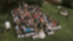 Bride and groom with a crowd of wedding guests outdoors, all facing the camera. Lush green lawn, white chairs, festive atmosphere.