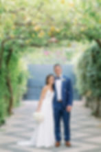 Bride in white gown and groom in blue suit stand under a lemon tree archway. She holds a bouquet. Greenery surrounds them, setting a romantic mood. Sorrento, Italy.