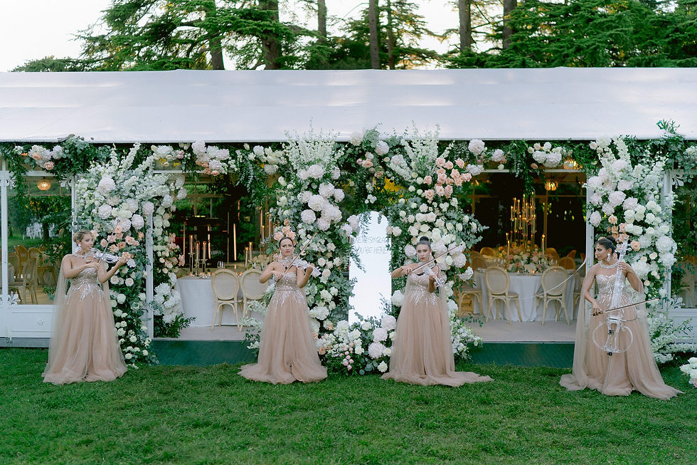 Four women in peach gowns play string instruments in front of a flower-adorned tent; elegant event setting with white and pastel florals. Lake Grada, Italy.