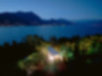 A lit tent in a lush garden by a lake at dusk. Mountains and city lights are visible across the water, creating a serene, scenic view. Lago Del Garda, Italy