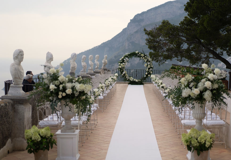 Outdoor wedding aisle with white floral arrangements, Roman bust statues, and a mountain view. Overcast sky sets a serene mood. Villa Cimbrone, Ravello.