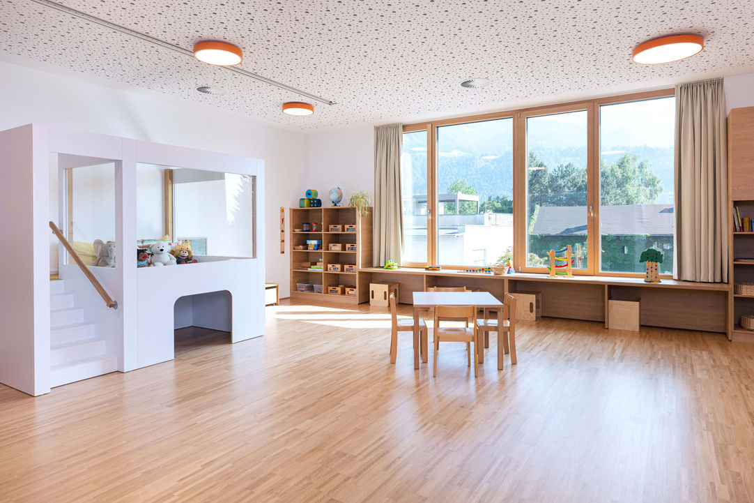 Gruppenraum mit Holzboden, Fensterband, sandfarbene Vorhänge, weißes Spielelement links, Holzregale, Tischgruppe zentral