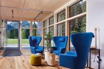 drei Designerstühle mit blauem Samtstoff, hölzerner Beistelltisch, im Hintergrund hölzerner, verglaster Eingangsbereich