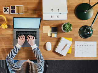 A clean, overhead shot of a home office surface — a shared calendar or planner open, a few sticky notes or cards nearby, and a phone or tablet showing a simple list or app. Everything looks organized, but there's no clear center of gravity.