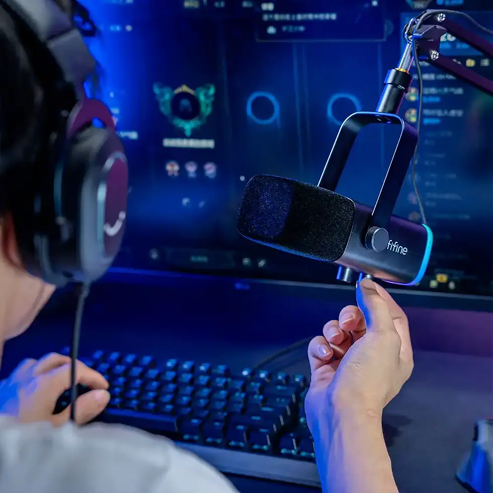 Person at a computer with headphones adjusts a microphone. A gaming screen glows blue in the background, creating a focused ambiance.