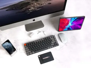 Laptop, mechanical keyboard with an orange key, and mouse on a white desk. Notepad with sketches and a plant nearby add freshness.