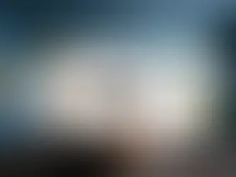 Person at a white desk with dual monitors displaying graphs. Holding a mug, surrounded by laptop, headphones, and stationery. Blue walls.