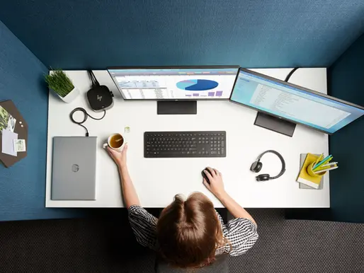 Person at a white desk with dual monitors displaying graphs. Holding a mug, surrounded by laptop, headphones, and stationery. Blue walls.