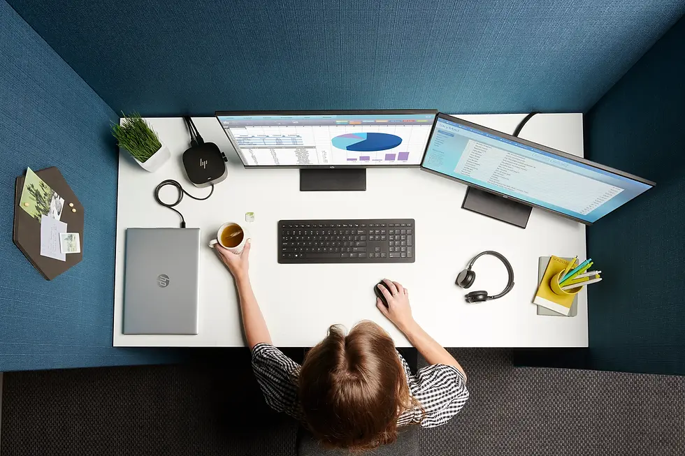 Person at a white desk with dual monitors displaying graphs. Holding a mug, surrounded by laptop, headphones, and stationery. Blue walls.