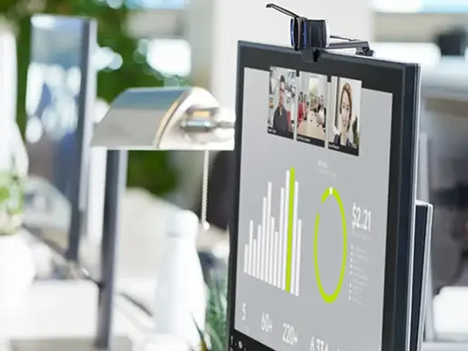 Computer screen in office shows a video call with charts and a $2.21 data point. Plant and lamp nearby, bright and modern setting.