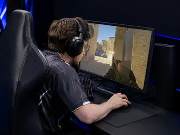 A person wearing headphones plays a shooting game on a large monitor. The setting is dark, and the screen shows a desert scene.