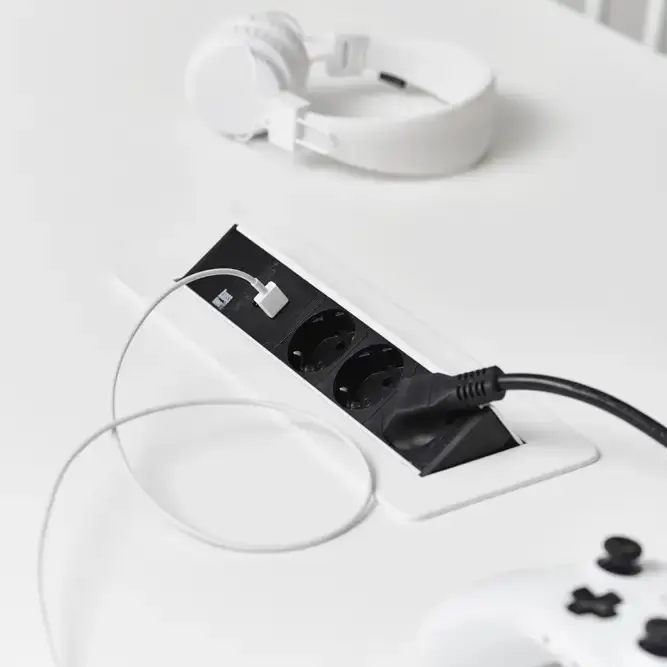 White desk with a built-in black power outlet, charging cables, white headphones, and a white game controller. Minimalist setup.