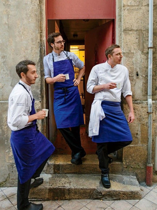 Three chefs in white jackets and blue aprons take a coffee break in a restaurant doorway.