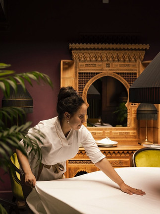 A housekeeper smooths a white cloth while setting a table in an elegant room.