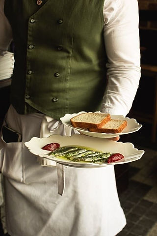 A waiter in a green waistcoat carries two plated dishes to a table.