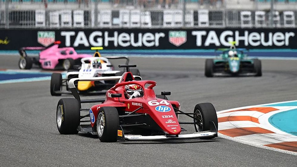 Red race car number 64 leads on a track, followed by pink and green cars. TAG Heuer ads line the background. High-speed action.