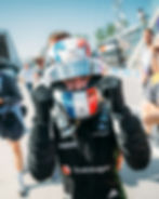 Racer in a colorful helmet and black suit celebrates with clenched fists on a sunny racetrack. Sponsors' logos are visible. Excited mood.