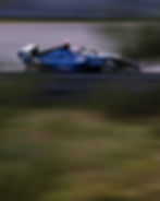 Blue race car number 57 speeds down a track, blurred motion. Sponsor logos visible. Greenery and barriers in the background.