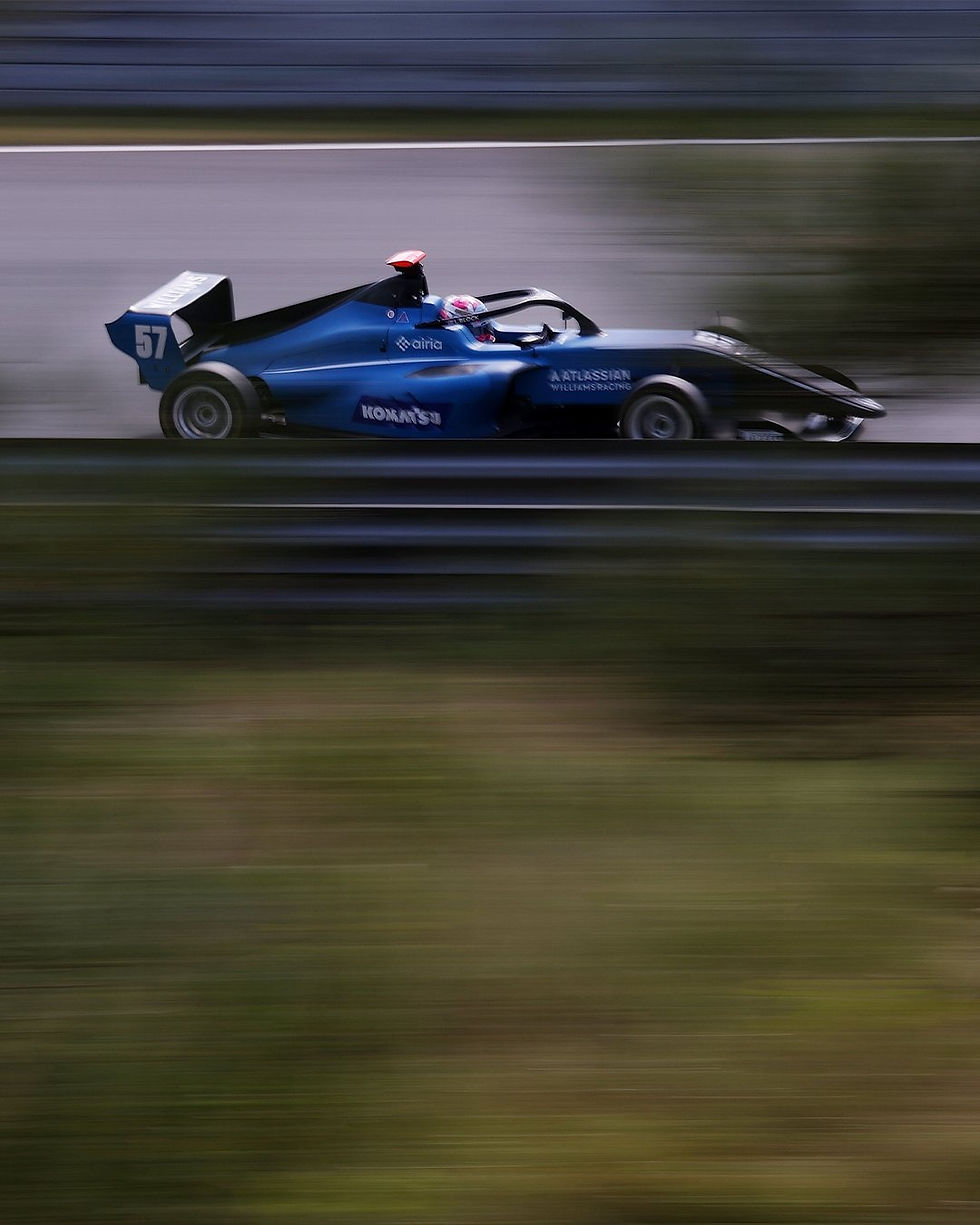 Blue race car number 57 speeds down a track, blurred motion. Sponsor logos visible. Greenery and barriers in the background.