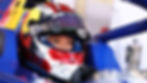 Racer in helmet with Red Bull logo, focused eyes visible. Helmet features stars and name "Chloe". Blue car in background.