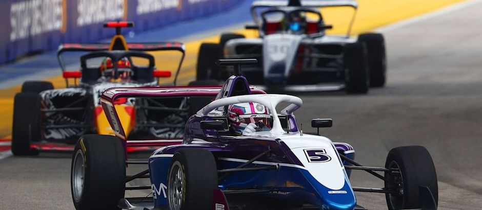 Racing cars on track with vibrant red, yellow, and purple designs. Car number 5 leads. Blue and yellow barriers line the course. Energetic mood.