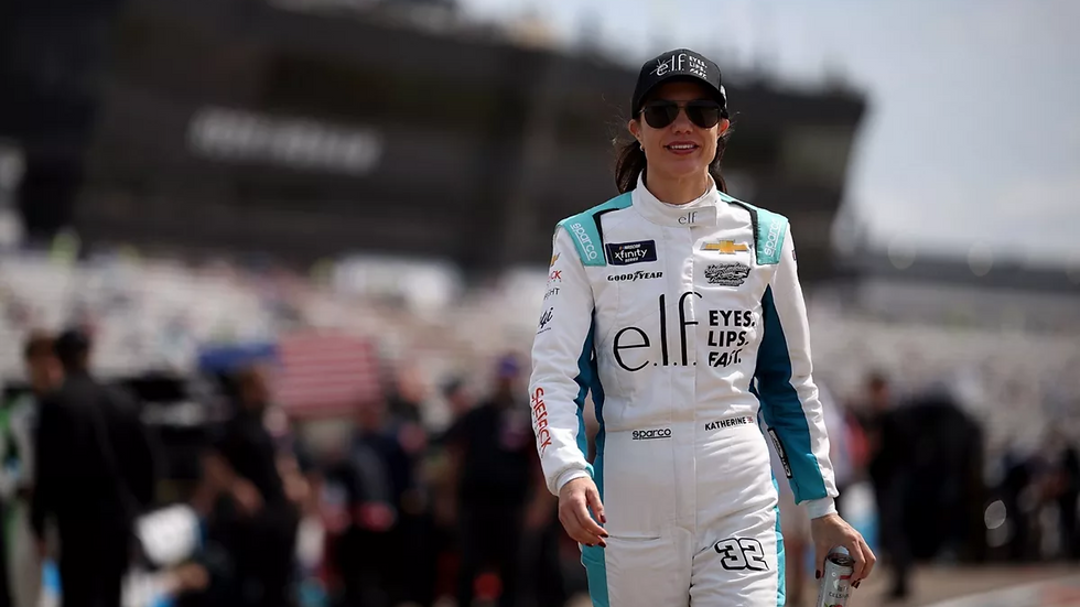 Race car driver in white and teal suit, wearing sunglasses and a cap, holds a can. Blurred racetrack background, sunny day.