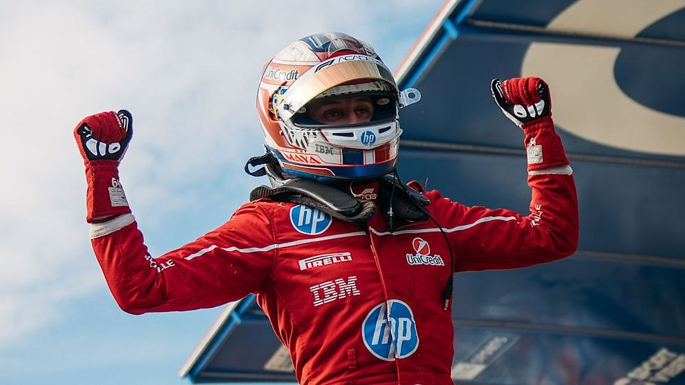 Racecar driver in a red suit with sponsor logos celebrates with raised fists, wearing a helmet. Blue sky and racing car wing in background.