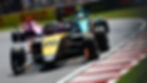 Race cars on a track, led by an orange and black car labeled 20. Pink and turquoise cars follow. Sunny day, green grass in background.