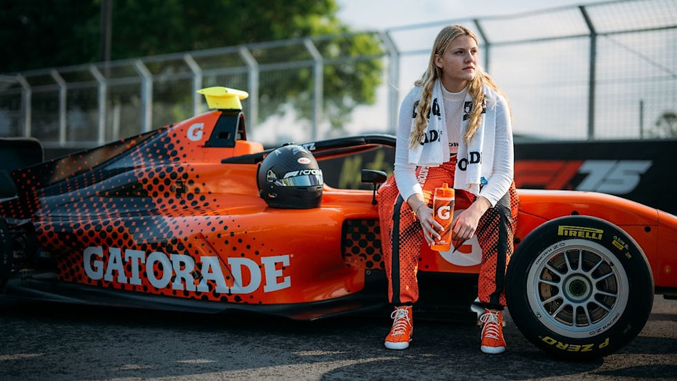Woman in racing suit sits on a bright orange Gatorade race car, holding a Gatorade bottle on a racetrack. Fence and trees in the background.