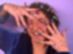 Katie Hornsby poses in a blue velvet top, her face is decorated with colorful nail stickers and nail art, smiling playfully against a purple background.
