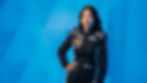 A person in a racing suit poses confidently against a blue geometric background. The suit displays several sponsor logos, showing a focused mood.