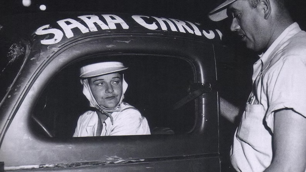 A woman in white racing gear sits in a car, labeled "Sara Christ" on the side. A man in a cap stands beside, leaning in at night.