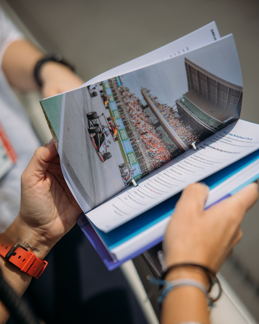 Hands hold an open magazine showing a racetrack photo with cars and spectators. A red watch is on the right wrist.