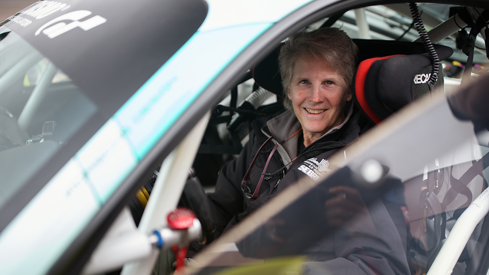 Smiling woman in racing gear sits in a sports car with visible roll cage. The seat is black with red accents. Text on jacket reads "SEBRING."